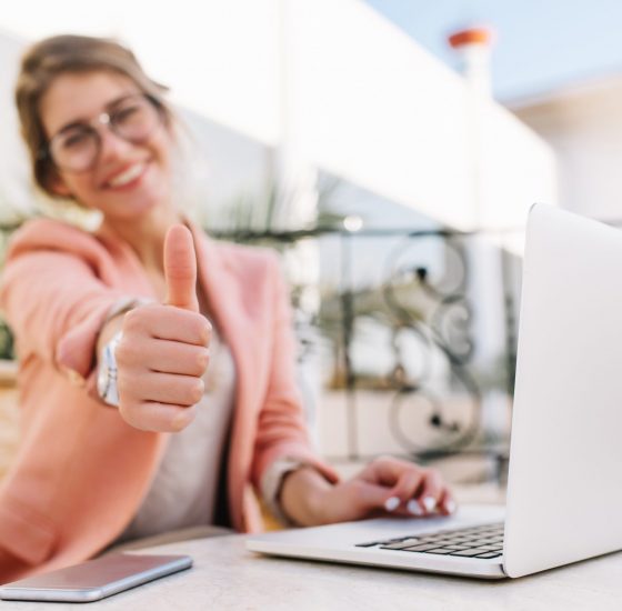 Cute young woman, student, business lady showing thumbs up, well done, sitting in outdoor cafe on terrace with laptop. Wearing pink smart clothes.